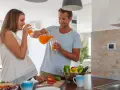 Szczęśliwy mężczyzna nalewa sok pomarańczowy ciężarnej kobiecie. W kuchni widać warzywa, co sugeruje zdrowe nawyki, ale pamiętaj o zakazanych sokach w ciąży.