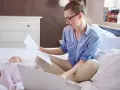 Kobieta w okularach siedzi na łóżku z laptopem i dokumentami, obok śpi dziecko. Załatwia formalności związane ze zmianą meldunku dziecka.