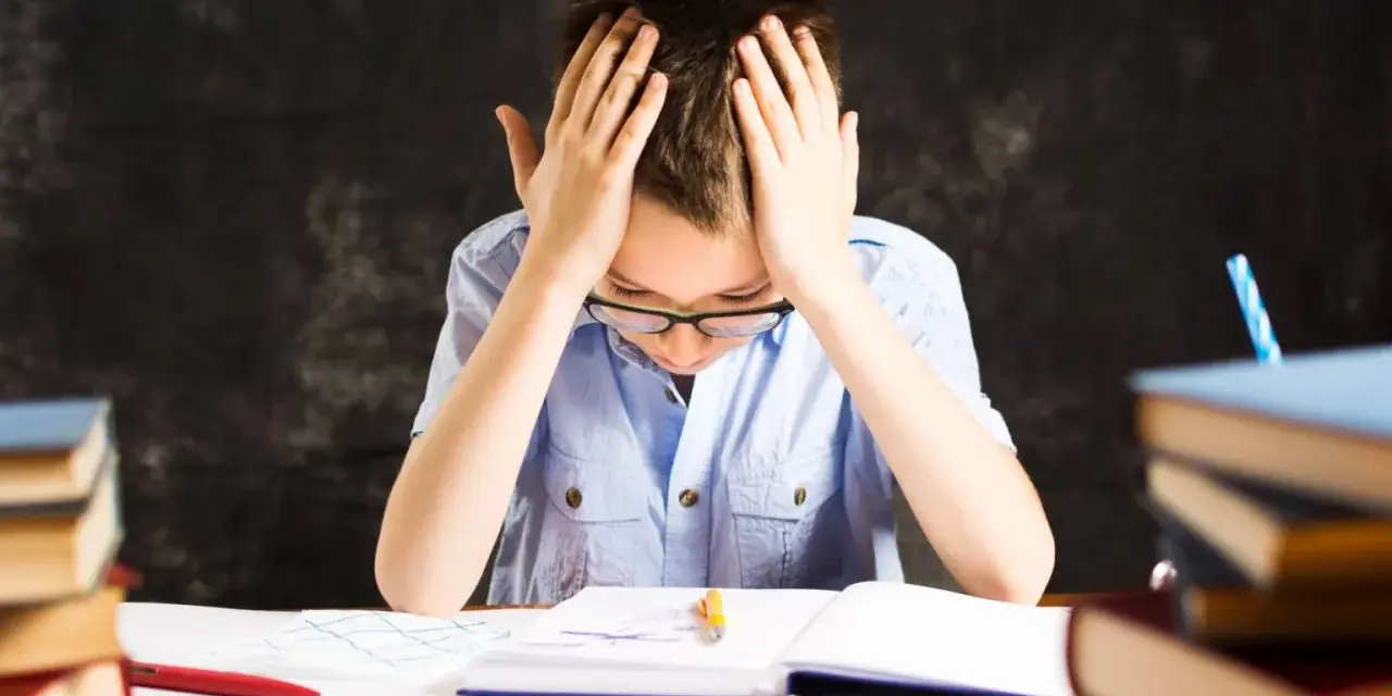 Chłopiec z rękami na głowie, otoczony książkami, zmaga się z nauką. Czy to dysleksja, co to za trudność?