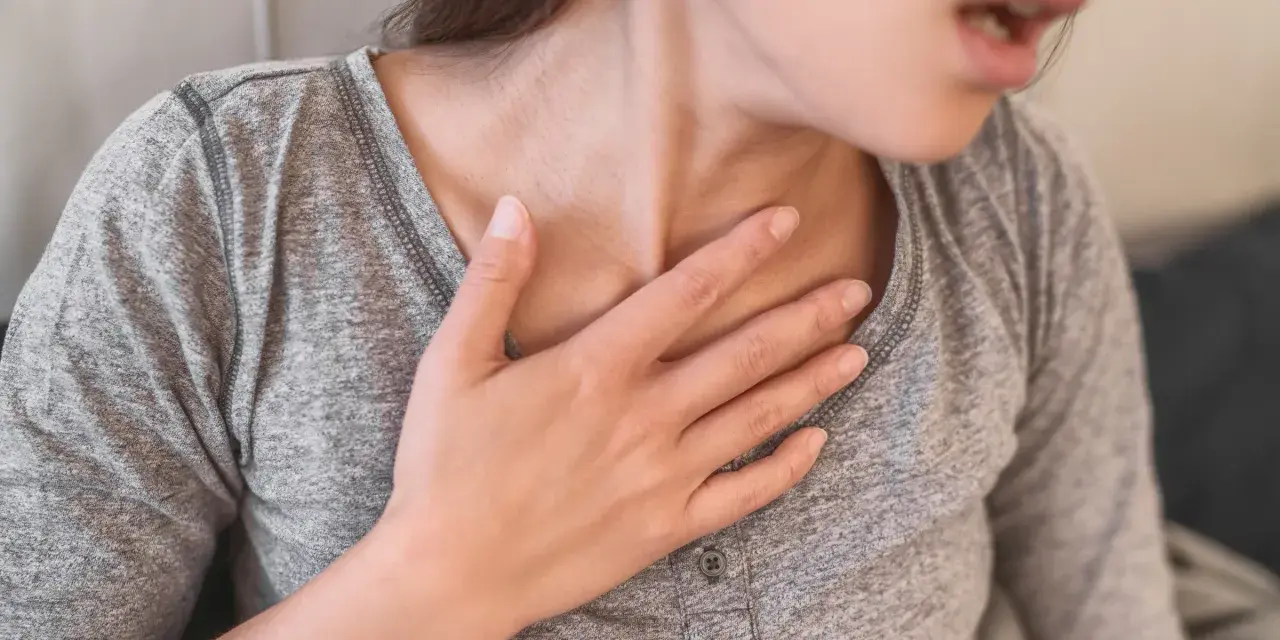 Kobieta z ręką na klatce piersiowej, odczuwająca duszności. To może być objaw po chorobie, wskazujący na potrzebę badań po covidzie.