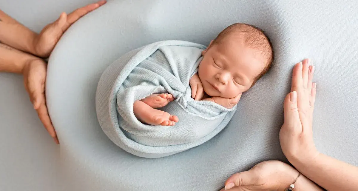 Otulony w błękitny kocyk śpi noworodek. Dookoła czuwają dłonie rodziców, tworząc bezpieczne gniazdko dla dziecka po 30. tygodniu ciąży.