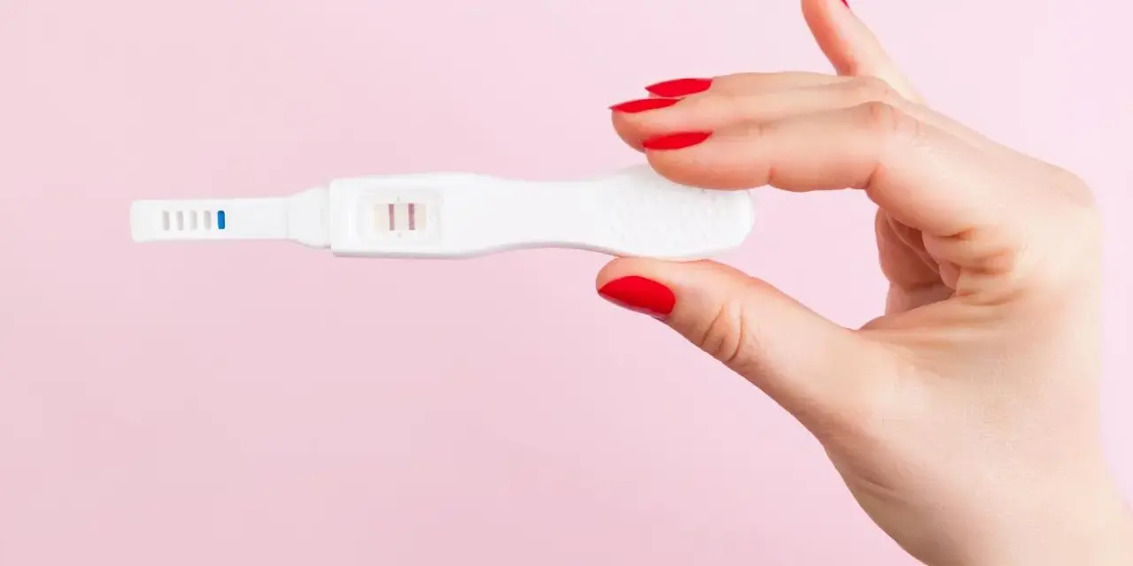 Dłoń z czerwonymi paznokciami trzyma test ciążowy z dwoma kreskami. Co może zafałszować wynik testu ciążowego?