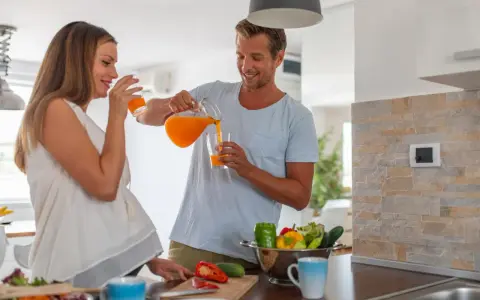 Szczęśliwy mężczyzna nalewa sok pomarańczowy ciężarnej kobiecie. W kuchni widać warzywa, co sugeruje zdrowe nawyki, ale pamiętaj o zakazanych sokach w ciąży.