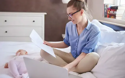 Kobieta w okularach siedzi na łóżku z laptopem i dokumentami, obok śpi dziecko. Załatwia formalności związane ze zmianą meldunku dziecka.