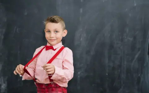 Chłopiec w różowej koszuli i czerwonych szelkach przed tablicą. Tak jak powinno czytać dziecko w 2 klasie, z uśmiechem i pewnością siebie.