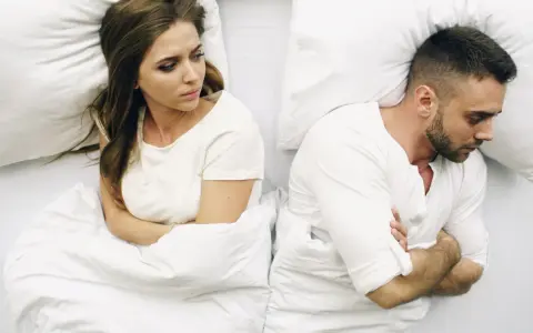 Para leży w łóżku odwrócona do siebie plecami. Kobieta patrzy zmartwiona, mężczyzna zamyślony. Może to być problem niskiego libido u mężczyzn.