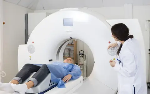 Pacjentka leży na stole tomografu, przygotowanie do tomografii z kontrastem. Lekarka obsługuje konsolę.