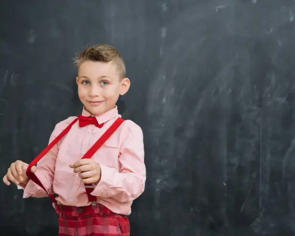 Chłopiec w różowej koszuli i czerwonych szelkach przed tablicą. Tak jak powinno czytać dziecko w 2 klasie, z uśmiechem i pewnością siebie.