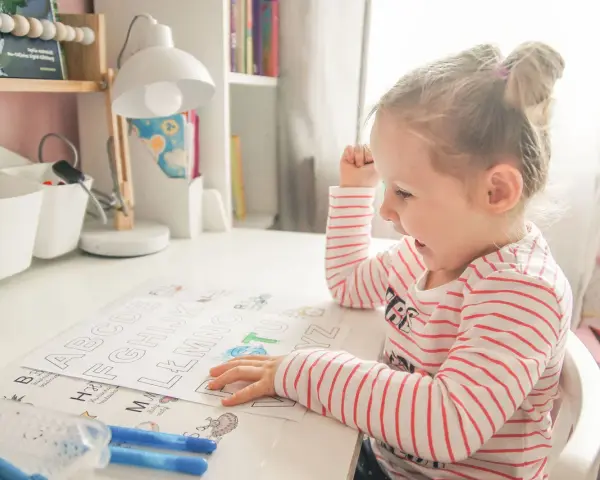Dziewczynka uczy się alfabetu na pamięć, kolorując litery. Zabawne obrazki pomagają w nauce.
