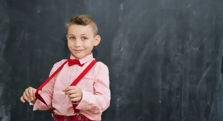 Chłopiec w różowej koszuli i czerwonych szelkach przed tablicą. Tak jak powinno czytać dziecko w 2 klasie, z uśmiechem i pewnością siebie.
