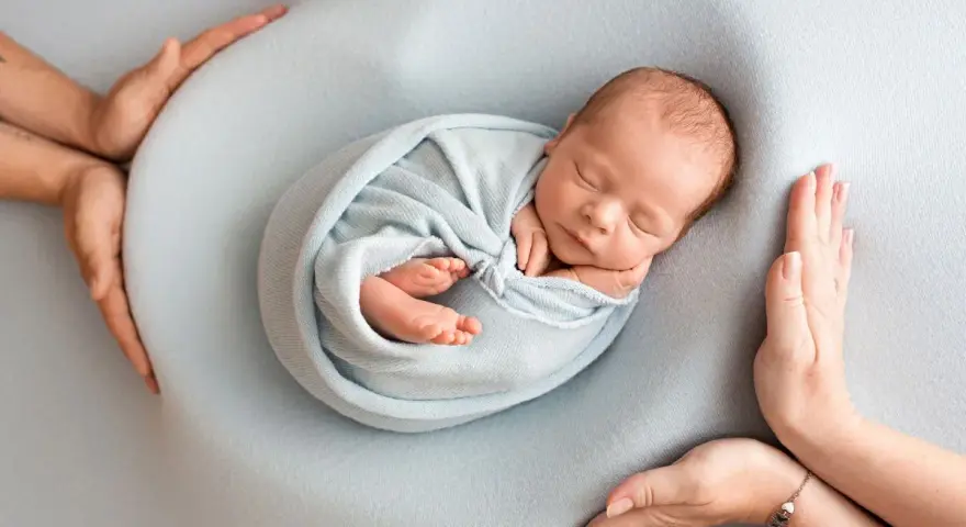Otulony w błękitny kocyk śpi noworodek. Dookoła czuwają dłonie rodziców, tworząc bezpieczne gniazdko dla dziecka po 30. tygodniu ciąży.