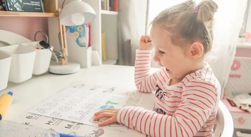 Dziewczynka uczy się alfabetu na pamięć, kolorując litery. Zabawne obrazki pomagają w nauce.