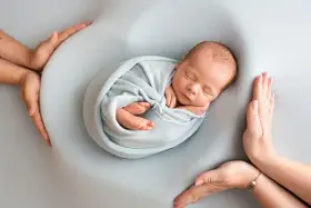 Otulony w błękitny kocyk śpi noworodek. Dookoła czuwają dłonie rodziców, tworząc bezpieczne gniazdko dla dziecka po 30. tygodniu ciąży.
