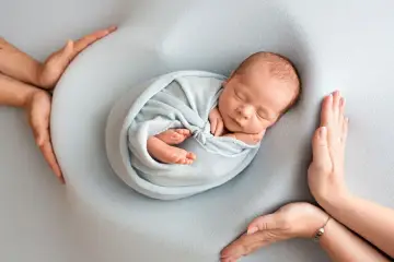 Otulony w błękitny kocyk śpi noworodek. Dookoła czuwają dłonie rodziców, tworząc bezpieczne gniazdko dla dziecka po 30. tygodniu ciąży.