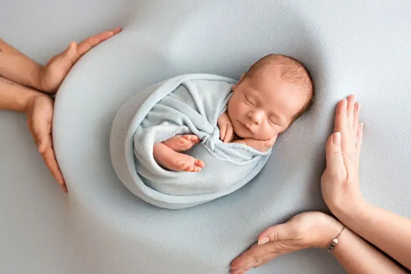 Otulony w błękitny kocyk śpi noworodek. Dookoła czuwają dłonie rodziców, tworząc bezpieczne gniazdko dla dziecka po 30. tygodniu ciąży.